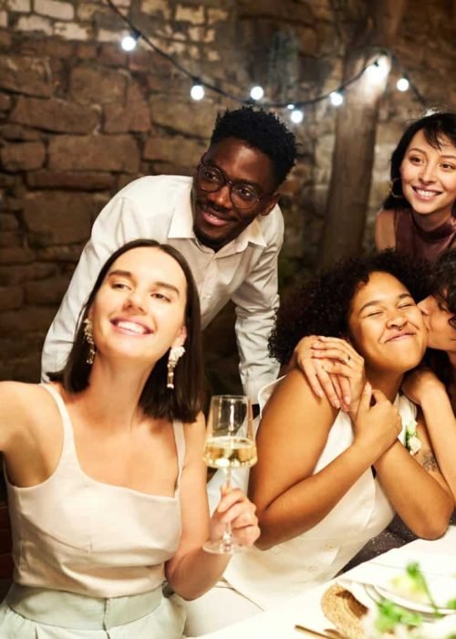 Young brunette woman taking selfie with group of friends and affectionate girl kissing her bride on cheek at wedding party